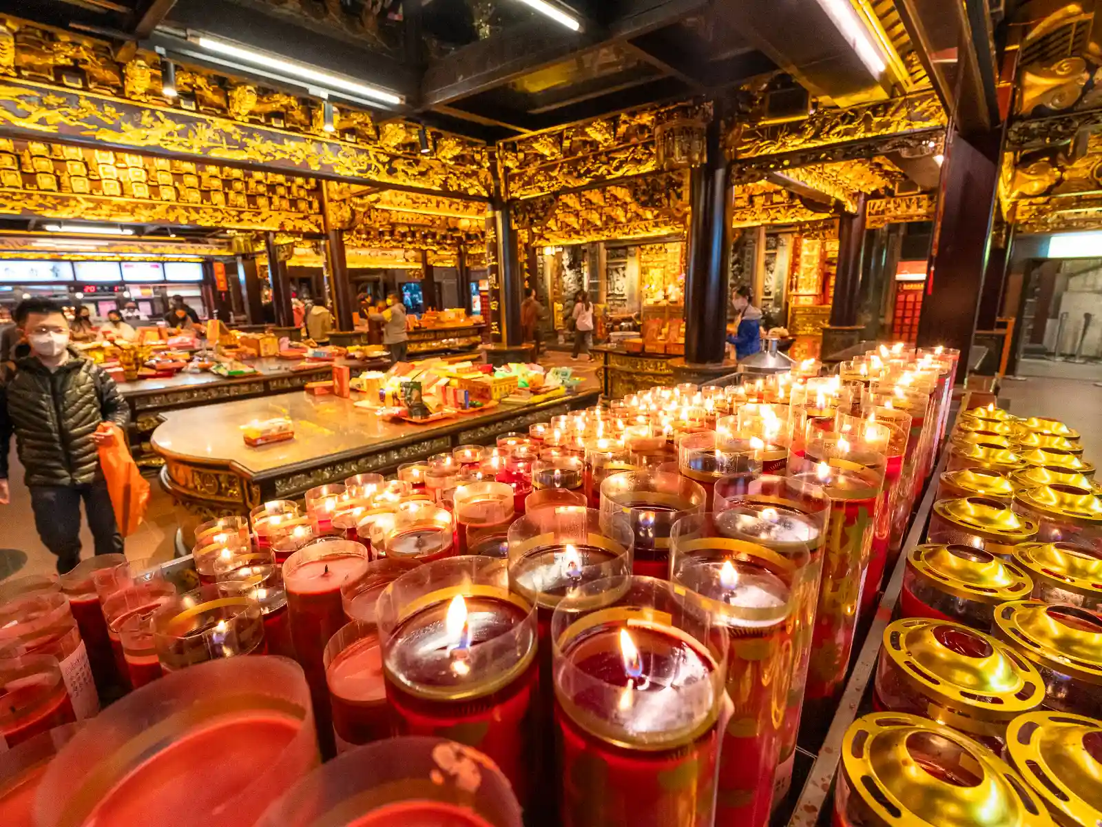 Rows of candles are burning on a table inside a room with a beautiful painted golden ceiling; sacrificial paper money is piled on adjacent tables.