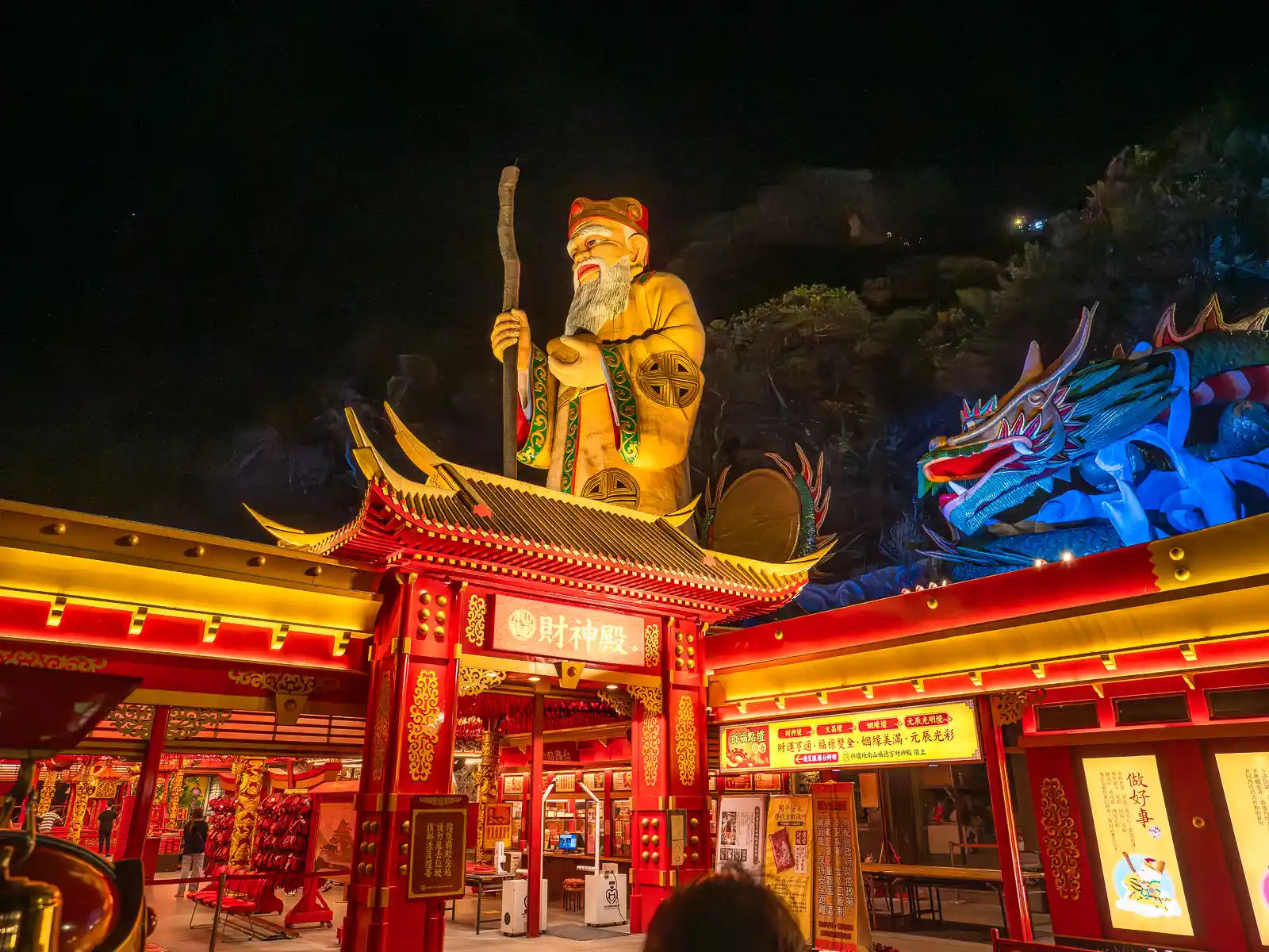 A giant Earth God statue, with a dragon on its side can be seen brightly illuminated on top of the God of Riches Temple at night.