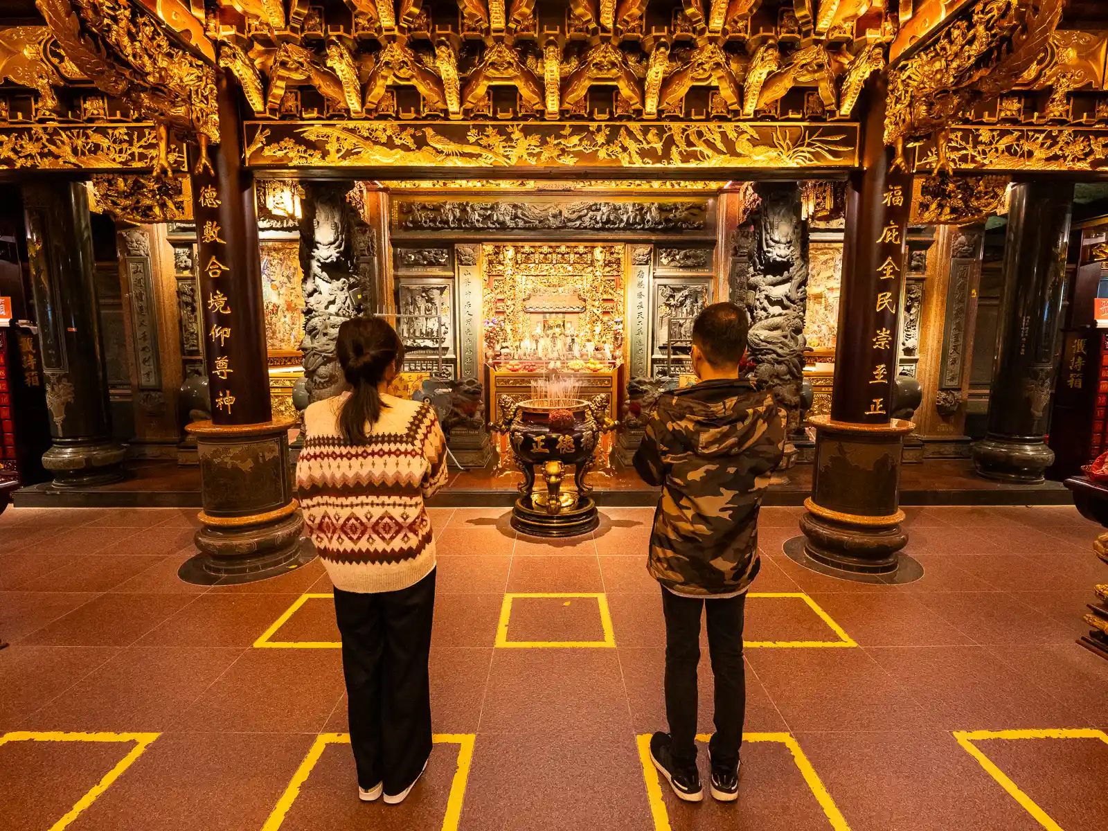 A couple prays in front of a wall decorated with beautiful painted wood carvings.