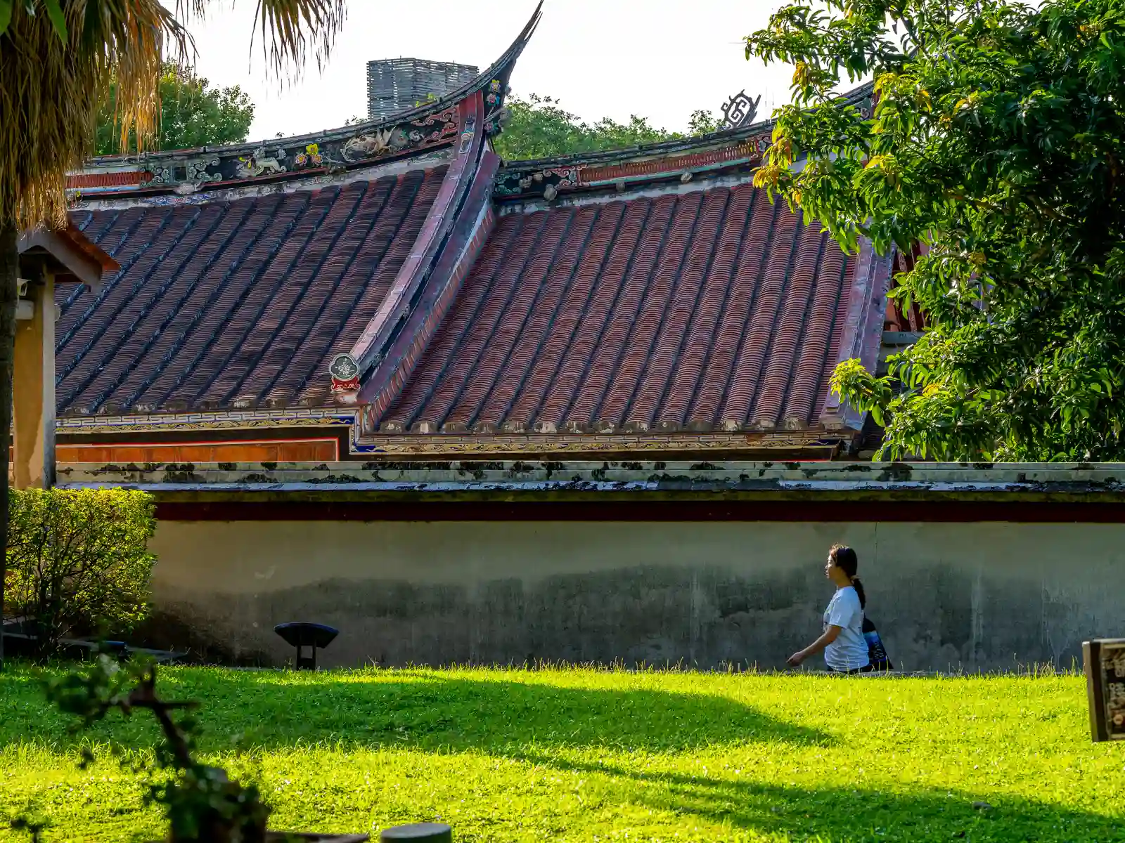 A tourist is seen walking in the gardens, above her, the roof ridge is decorated with painted wooden carvings.