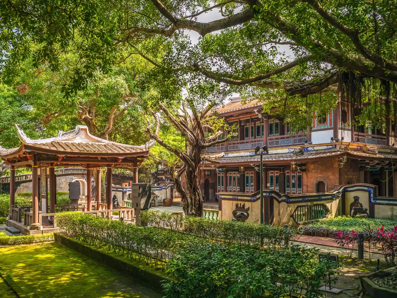 A beautiful wooden pavilion and one building of the Lin Family Mansion are visible between the plants of a lush and neatly-manicured garden.