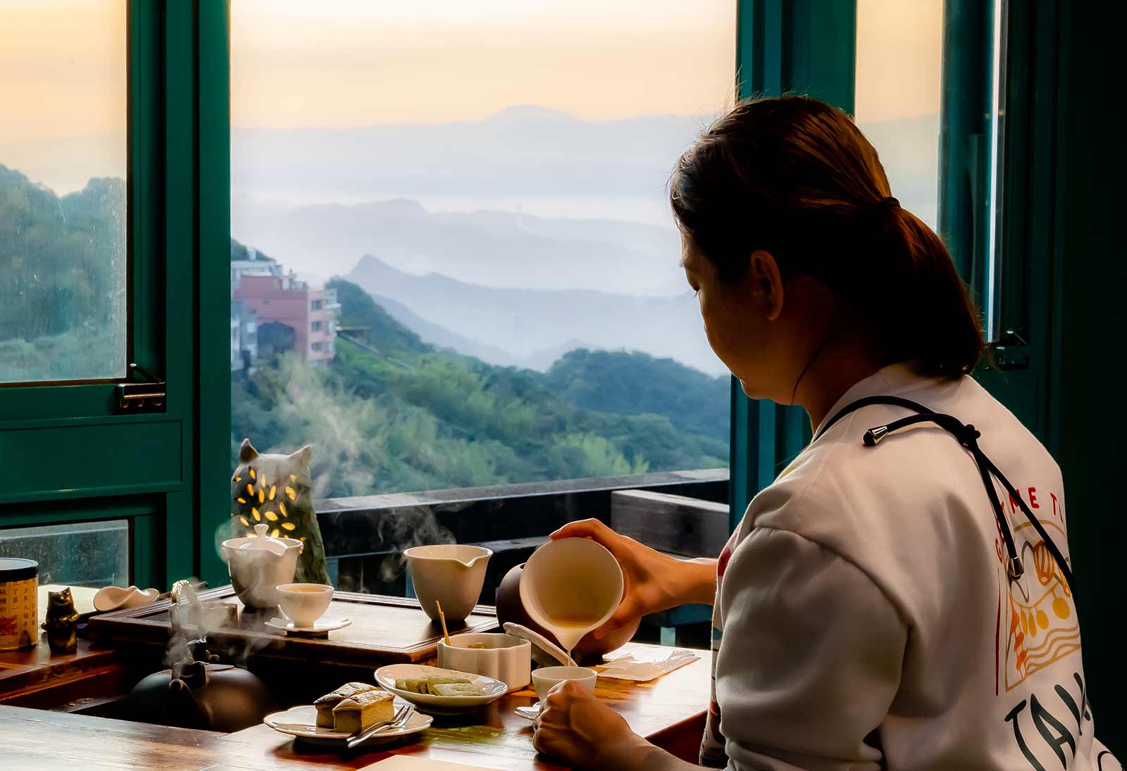 A guest makes tea in Jiufen Tea House with layers of mountains visible through the open window.