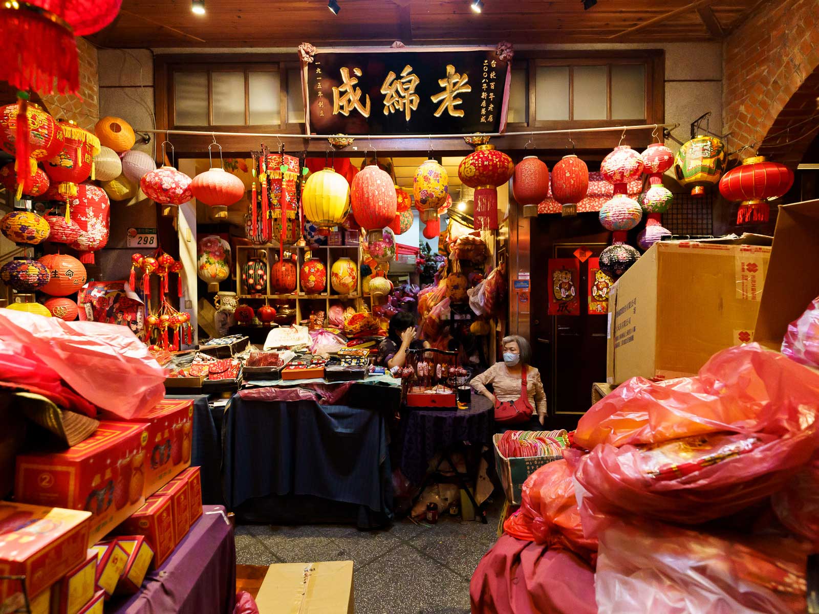 The walls of a storefront are decorated with red paper lanterns and boxes of Luner New Year's goods are piled around the room ready to be sold.