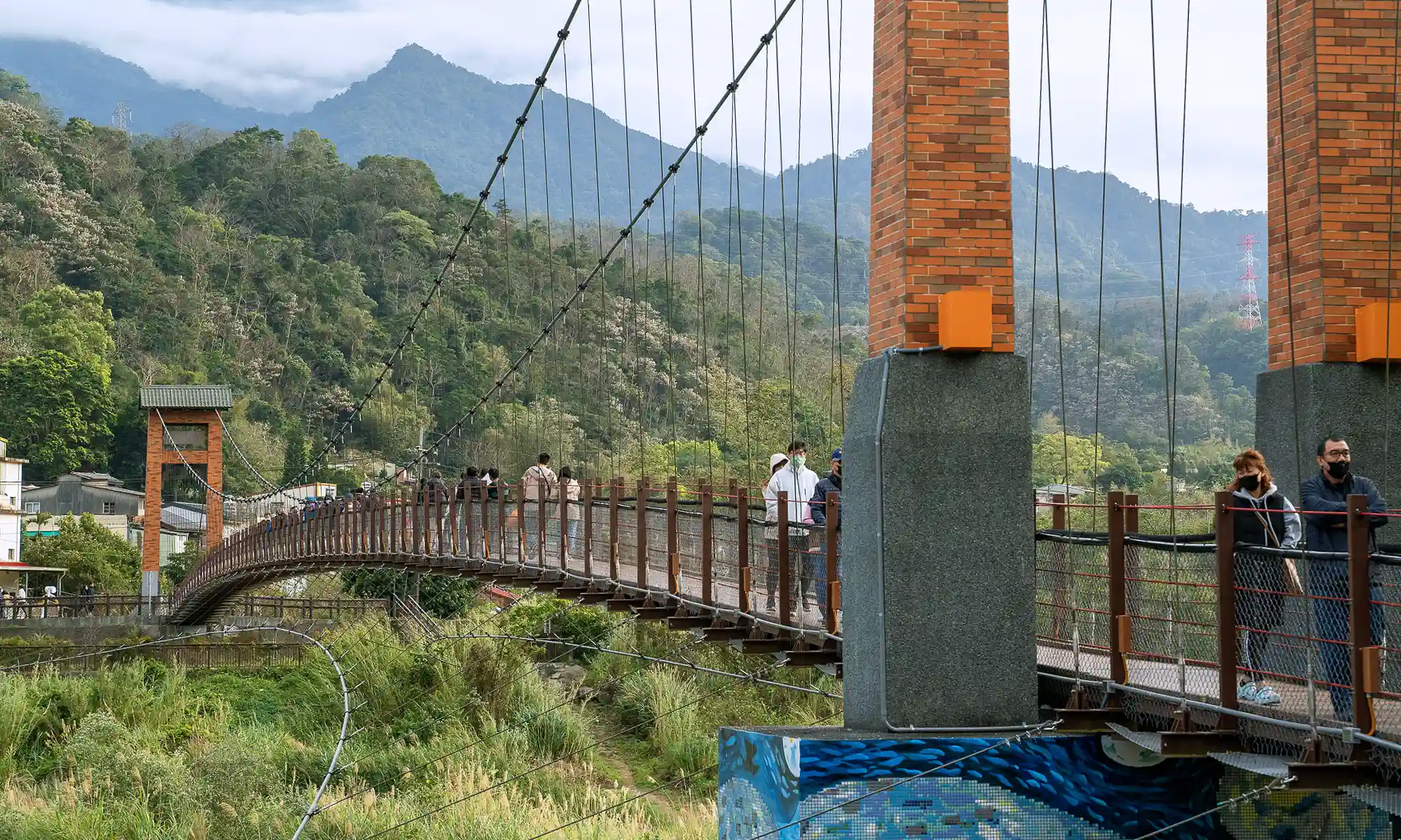 People walk over the Kangji Suspension Bridge; buildings and mountains can be seen in the distance.