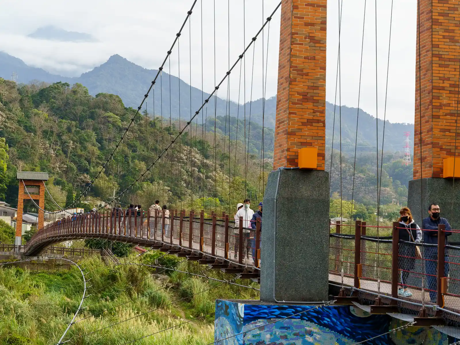 People walk over the Kangji Suspension Bridge; buildings and mountains can be seen in the distance.