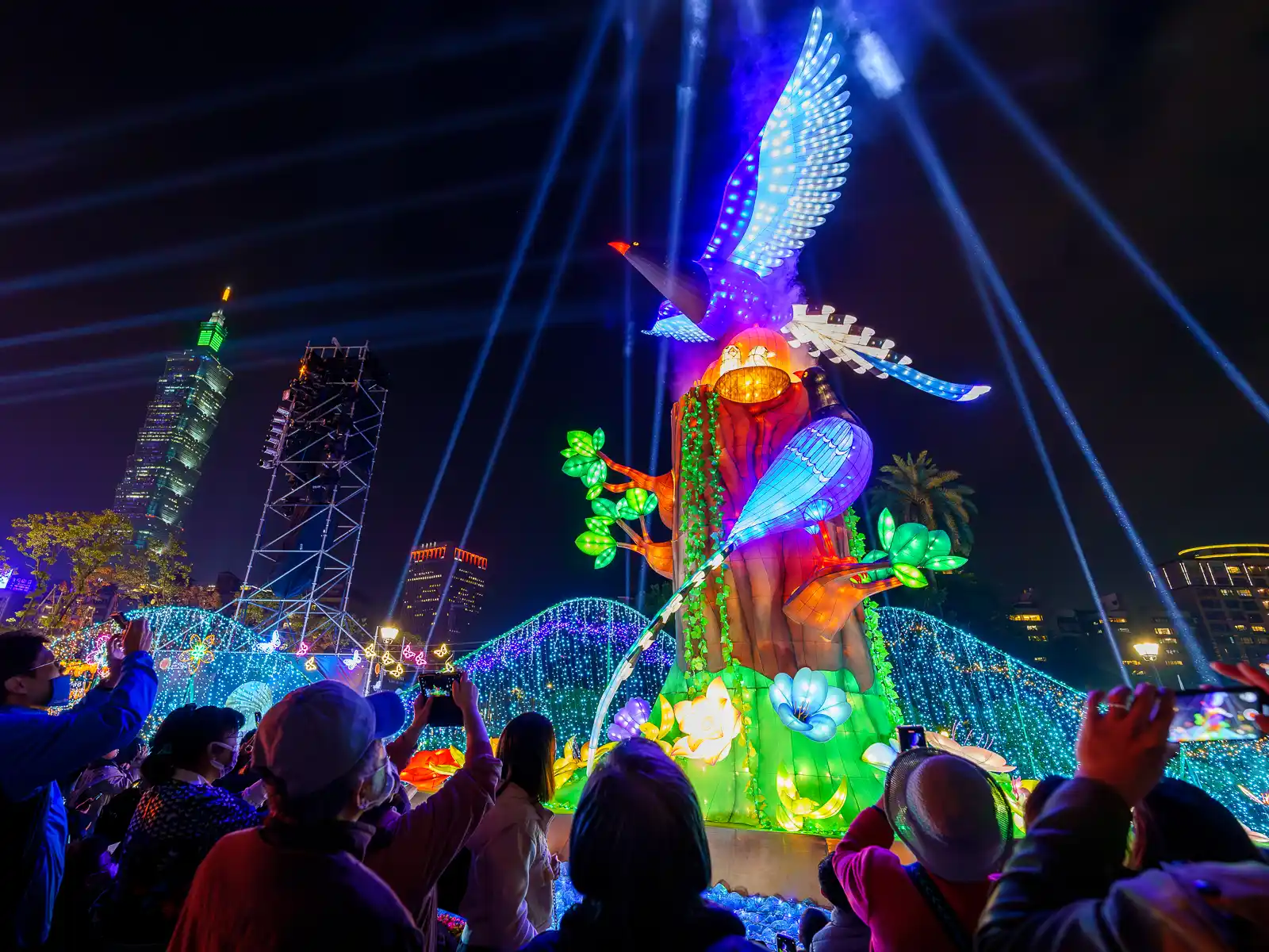 A tall mechanical lantern with a glowing bird towers above a crowd as a light show happens in the background.