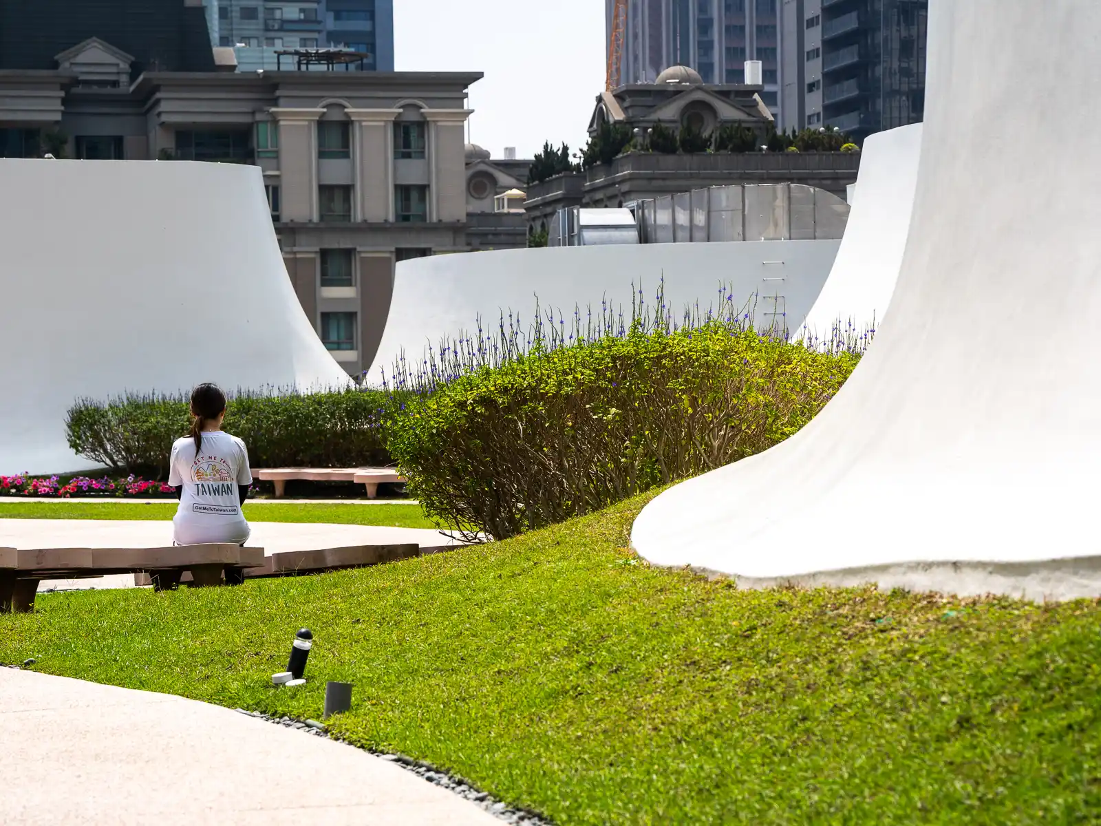 A tourist sits on a bench surrounded by large curved cement shapes at the National Taichung Theater's Rooftop Garden.