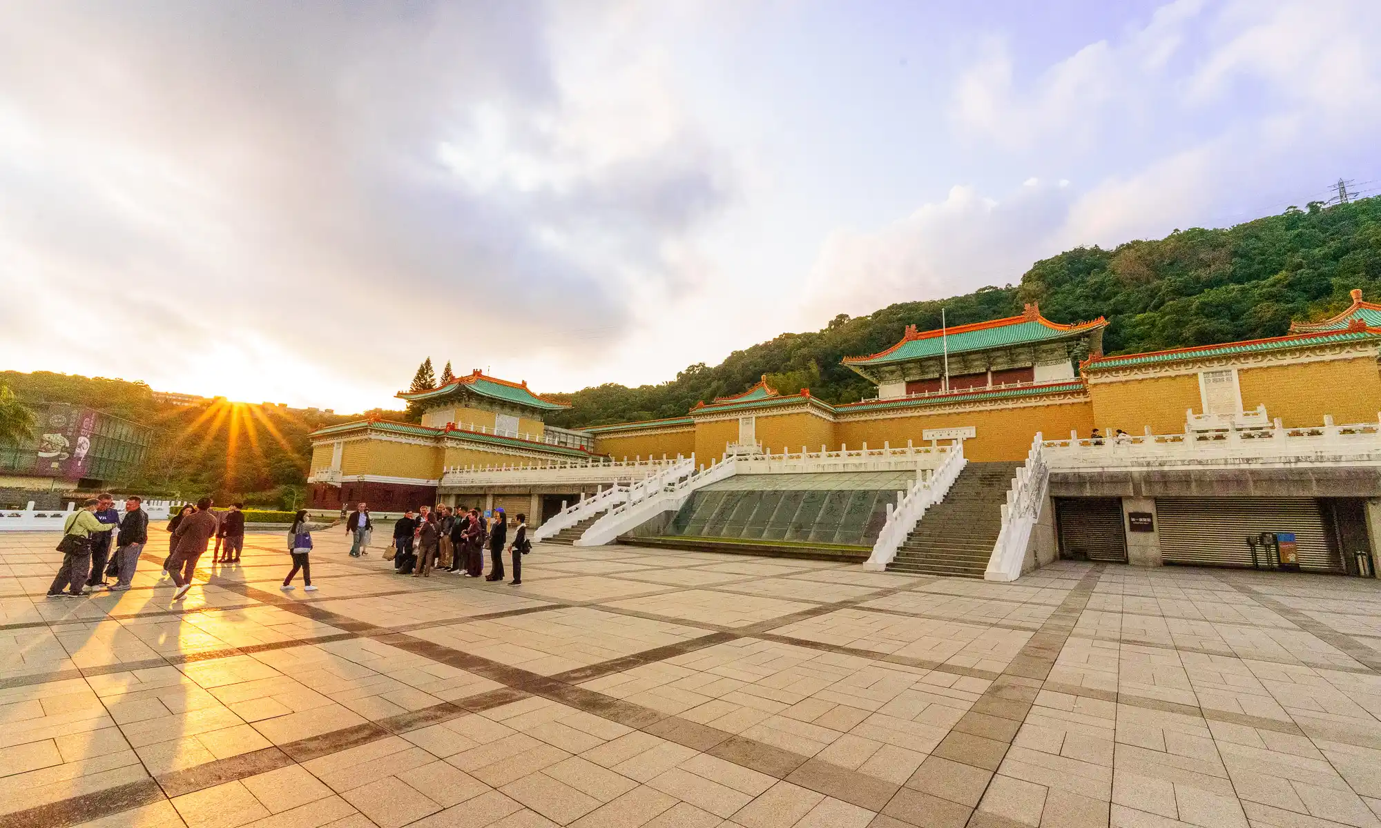 The sun sets behind the traditional architecture of the National Palace Museum while people walk through an expansive tiled plaza.